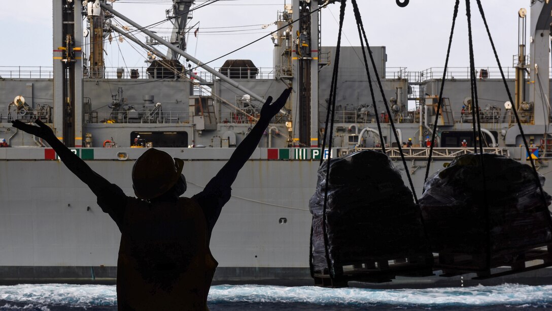 A sailor, shown in silhouette, extends her arms in a "Y" to signal from aboard one ship to another ship, which is sending cargo via cable.