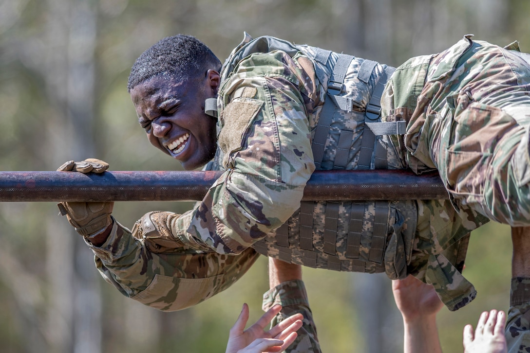 A soldier winces while holding onto a horizontal metal bar, as fellow troops' hands reach up to support him.