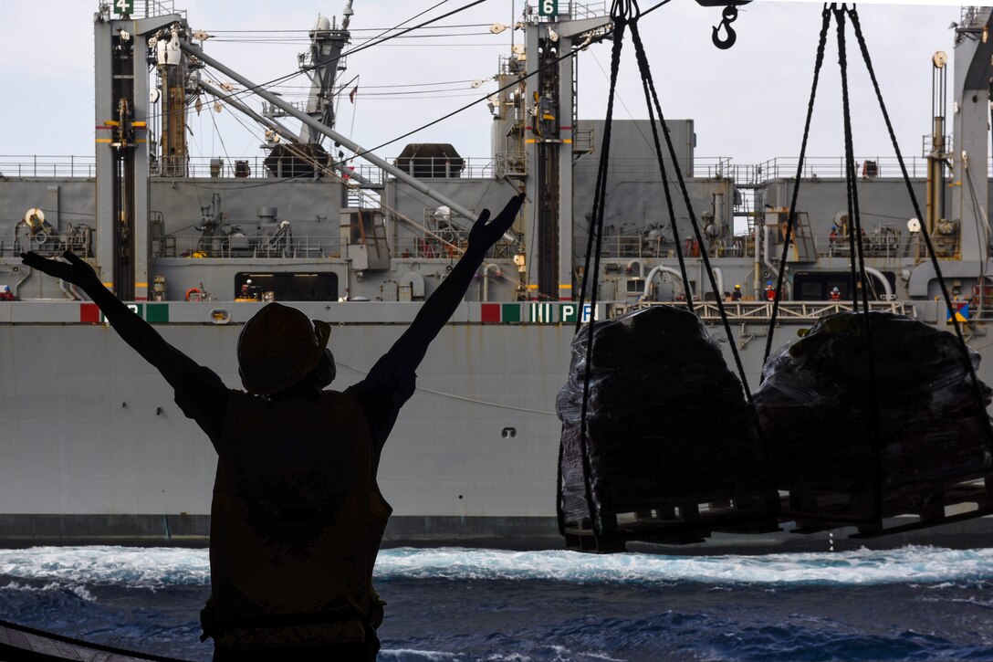 A sailor, shown in silhouette, extends her arms in a "Y" to signal from aboard one ship to another ship, which is sending cargo via cable.