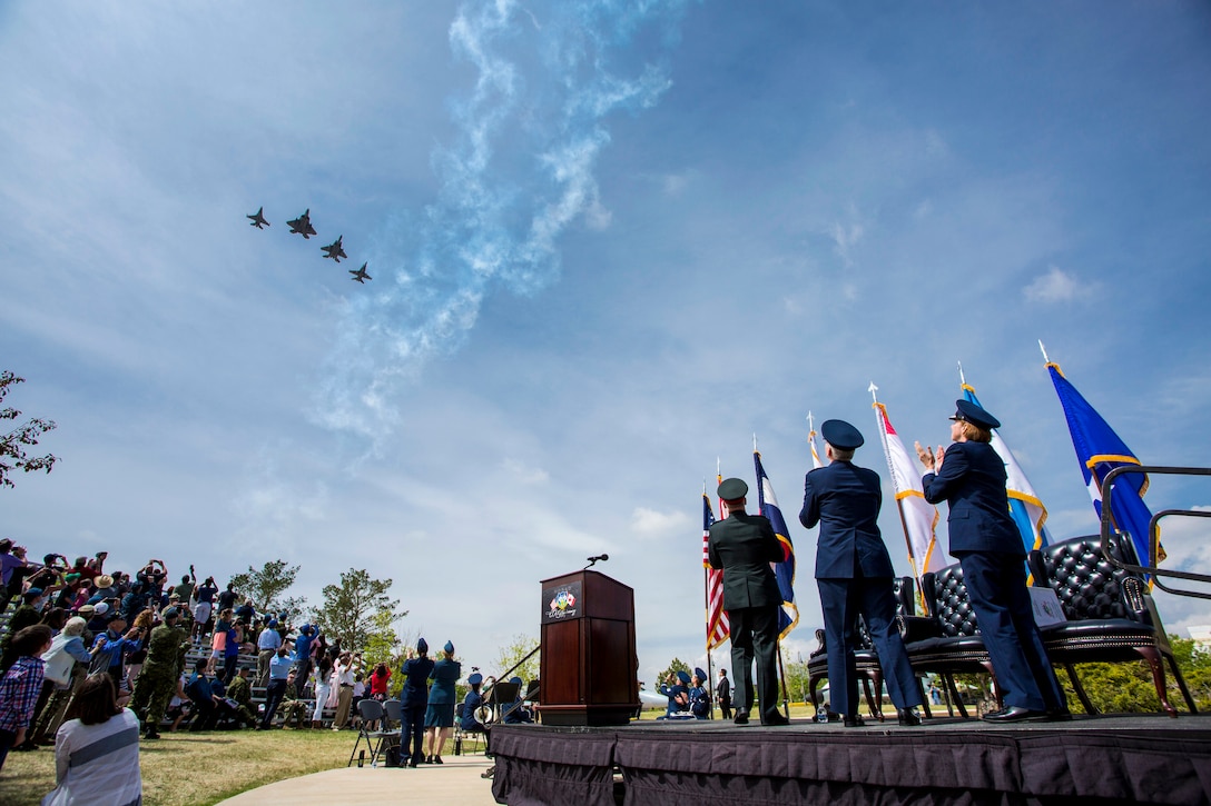 Four aircraft fly in formation as service members on a stage and crowd members observe.