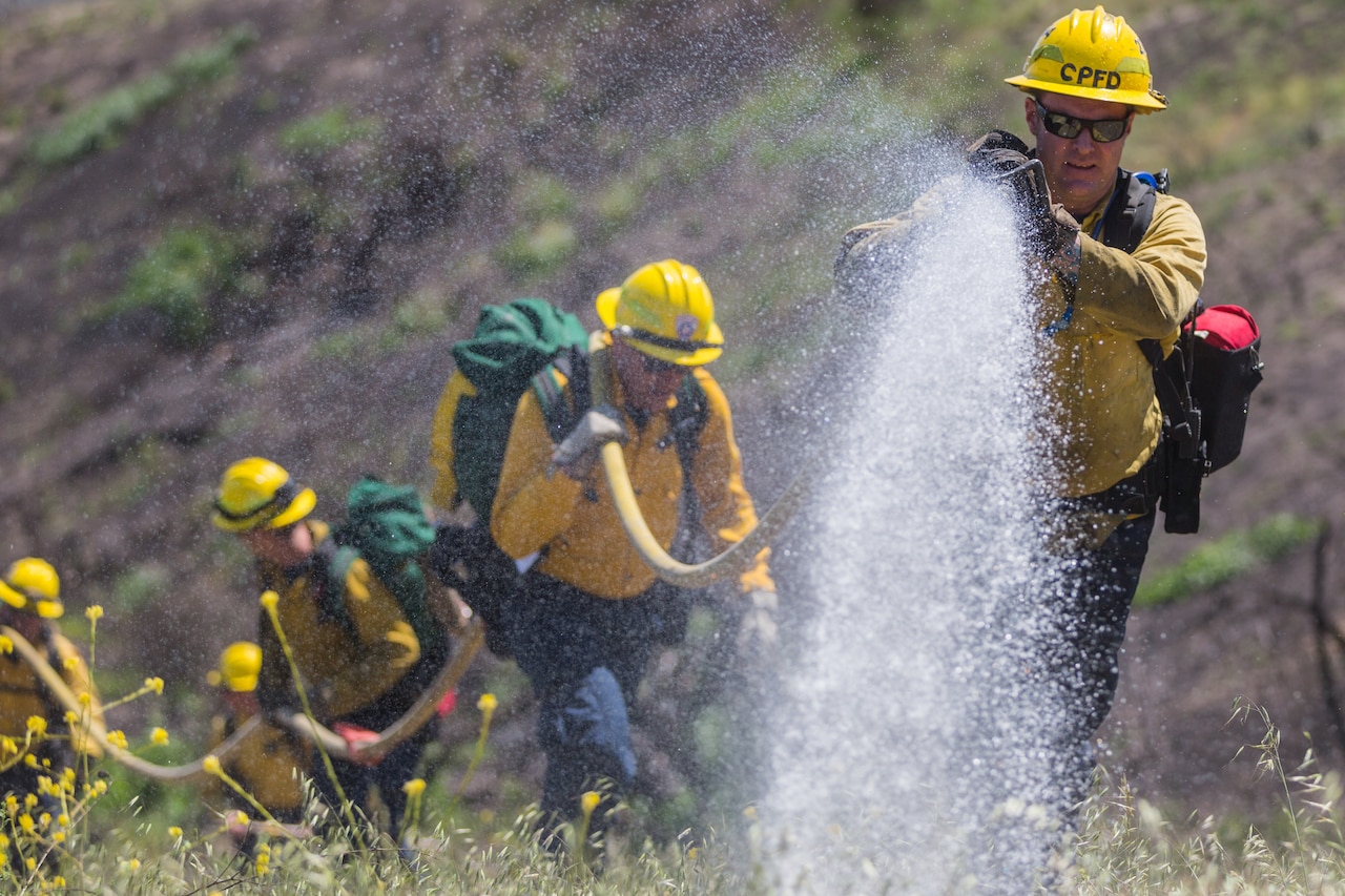 Firefighters carry a hose spouting water as they make their way up a grassy hill.