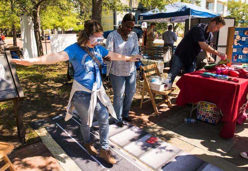 Carol Lampkinharris, Joint Base Charleston drug testing program administrative manager, assists an attendee wearing 'drunk goggles' during the fourth annual Federal Executive Association Government Expo May 11, 2018, at Liberty Square, Charleston, S.C.