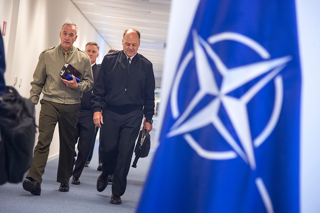 Two military leaders walk down a hallway with a NATO flag on the side.