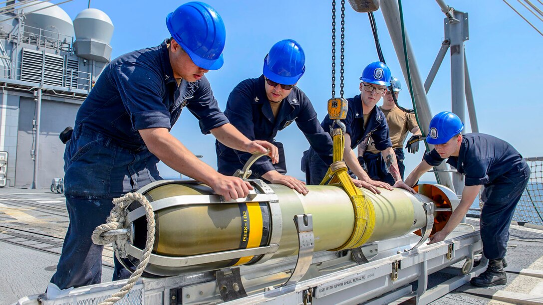 Four sailors in blue hardhats secure a torpedo to a platform on a ship's deck.