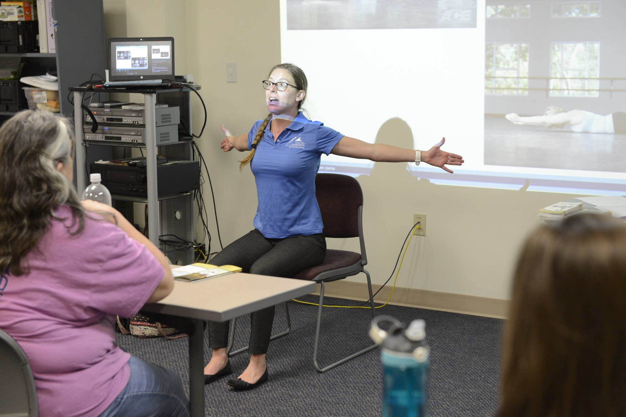 White female sitting in a chair with her arms stretched wide as she shows how to stretch out your back and shoulders.