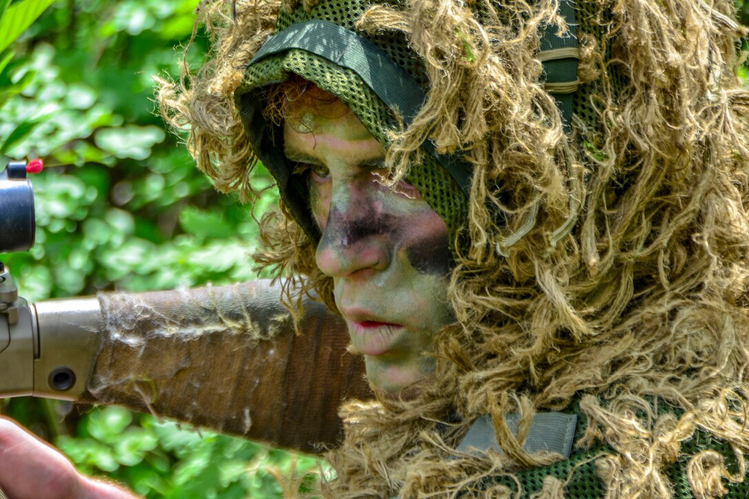 A sniper, in camouflage face paint and a head covering resembling vegetation, stares and points a weapon in a woodsy area.