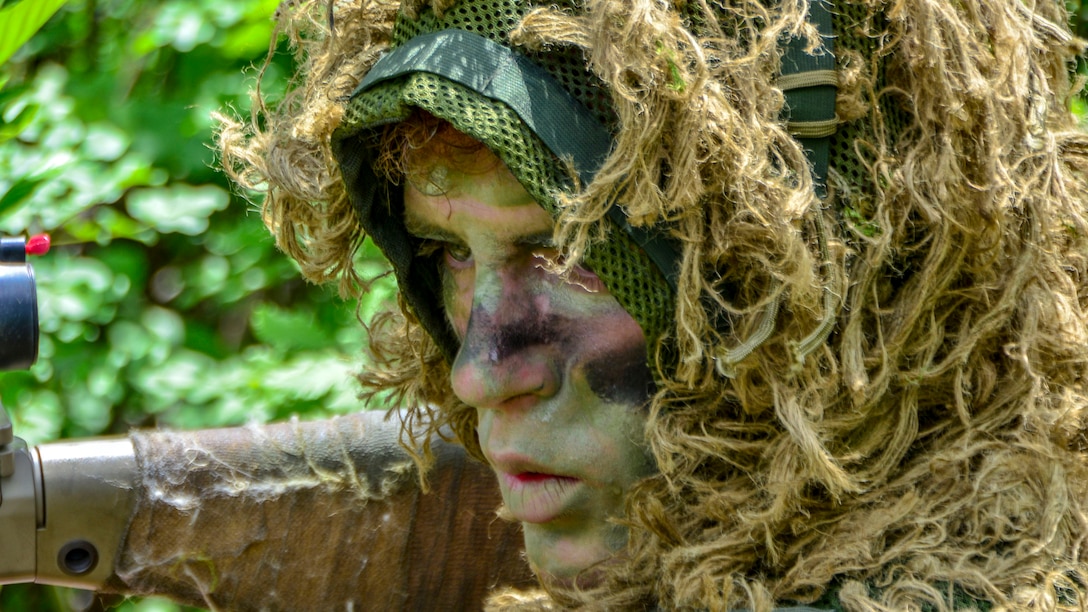 A sniper, in camouflage face paint and a head covering resembling vegetation, stares and points a weapon in a woodsy area.