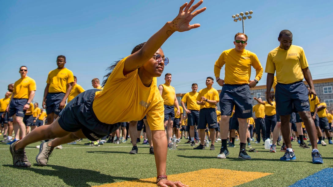 A sailor does a one-handed pushup on a field as others stand and watch.