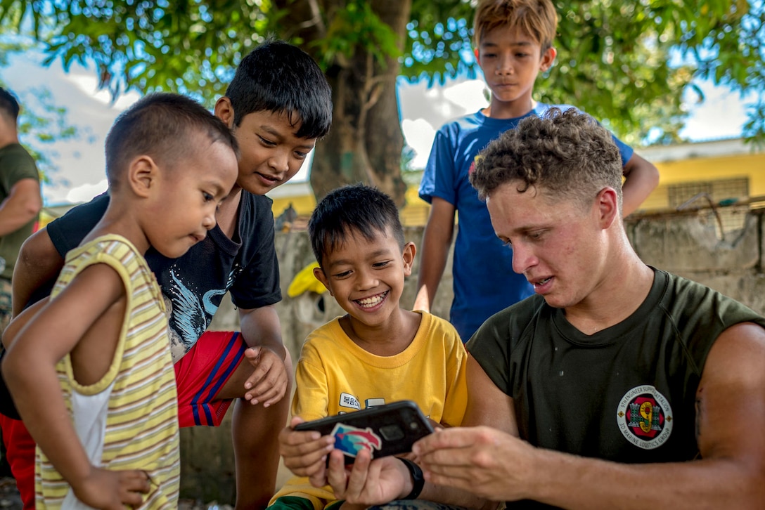 A Marine shows his phone to a group of boys while sitting outside.