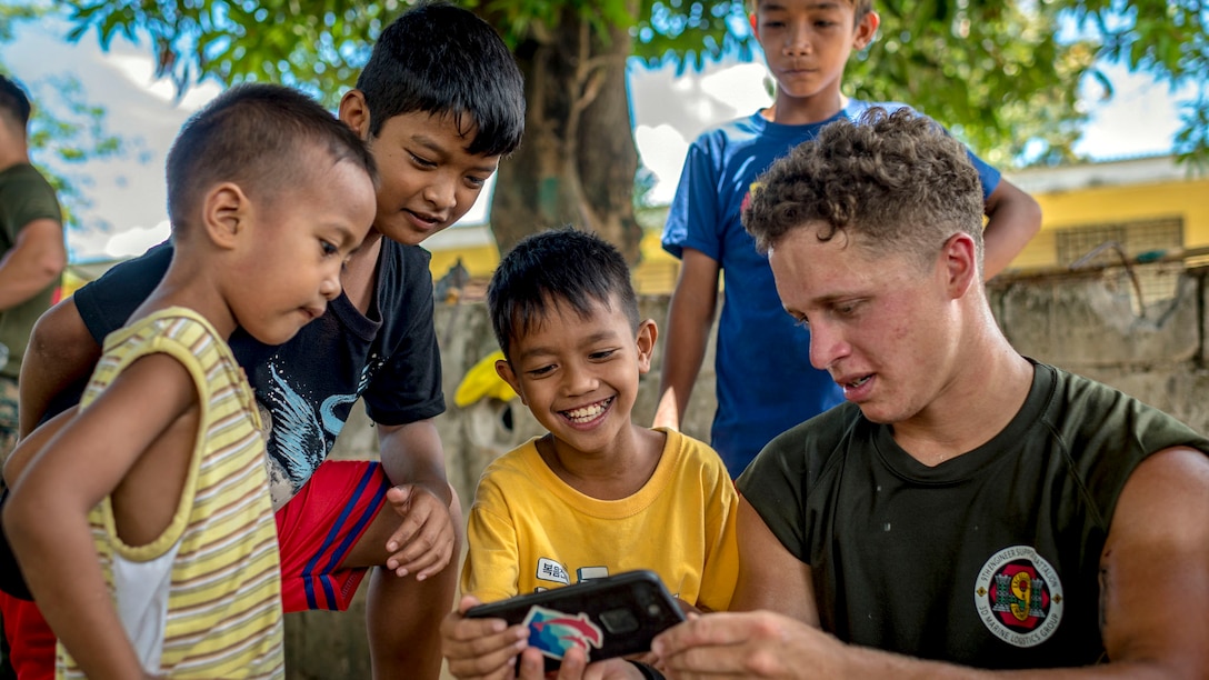 A Marine shows his phone to a group of boys while sitting outside.