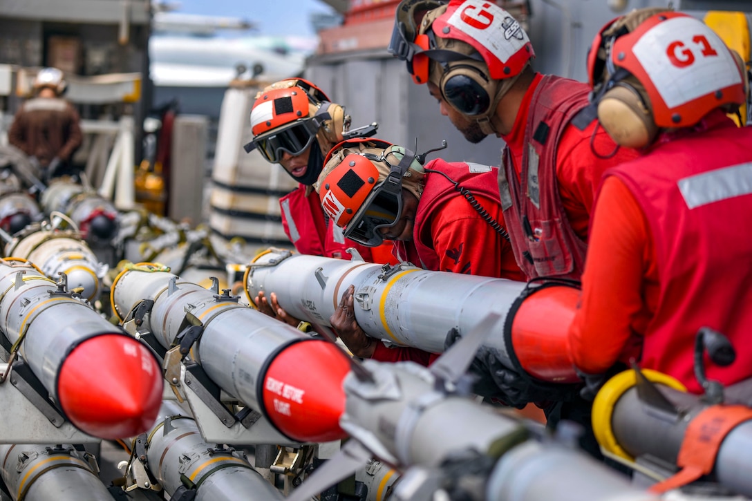 Sailors wearing red shirts and helmets move red-tipped missiles on a ship's flight deck.