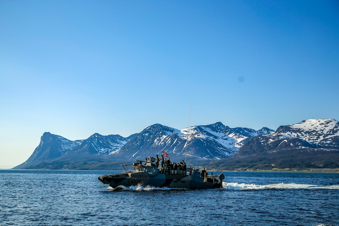 A fast assault craft travels in water with snow-capped mountains in the background.