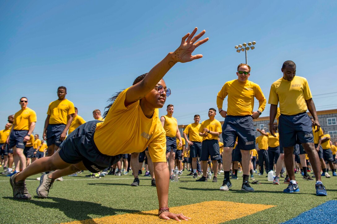 A sailor does a one-handed pushup on a field as others stand and watch.