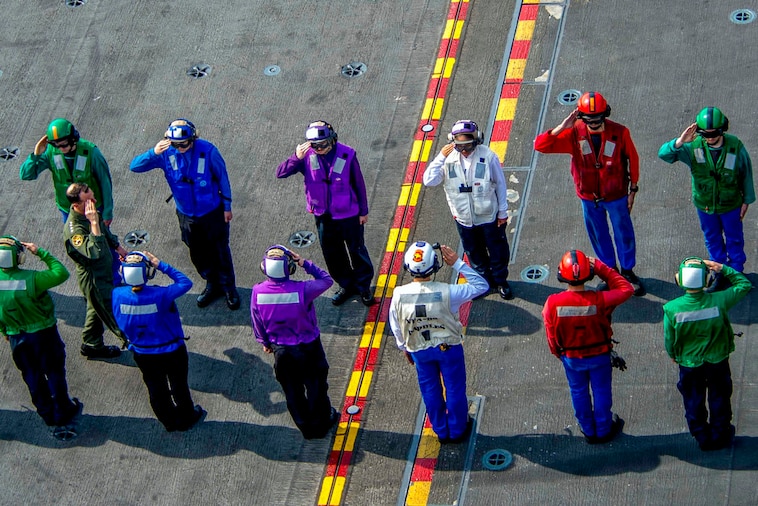 Two rows of sailors in uniforms of different primary colors salute.