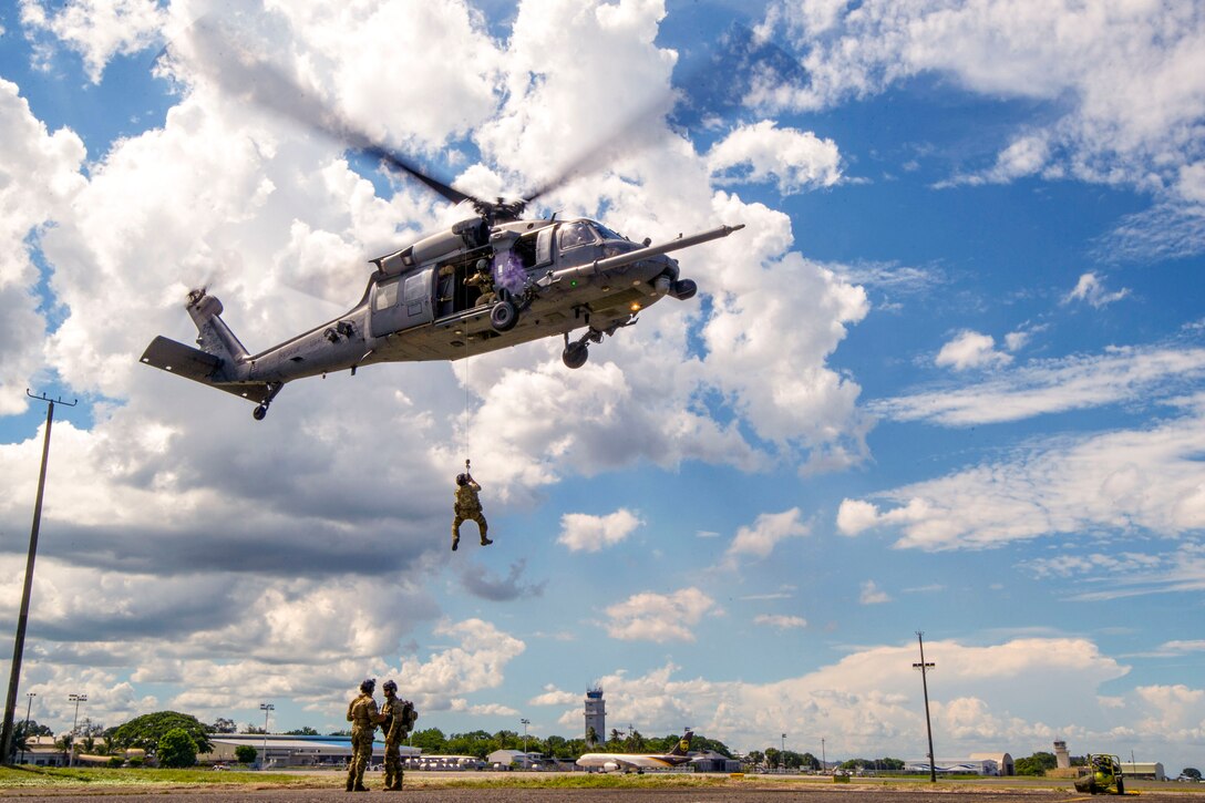An airman hangs from a helicopter by a line as people on the ground observe.