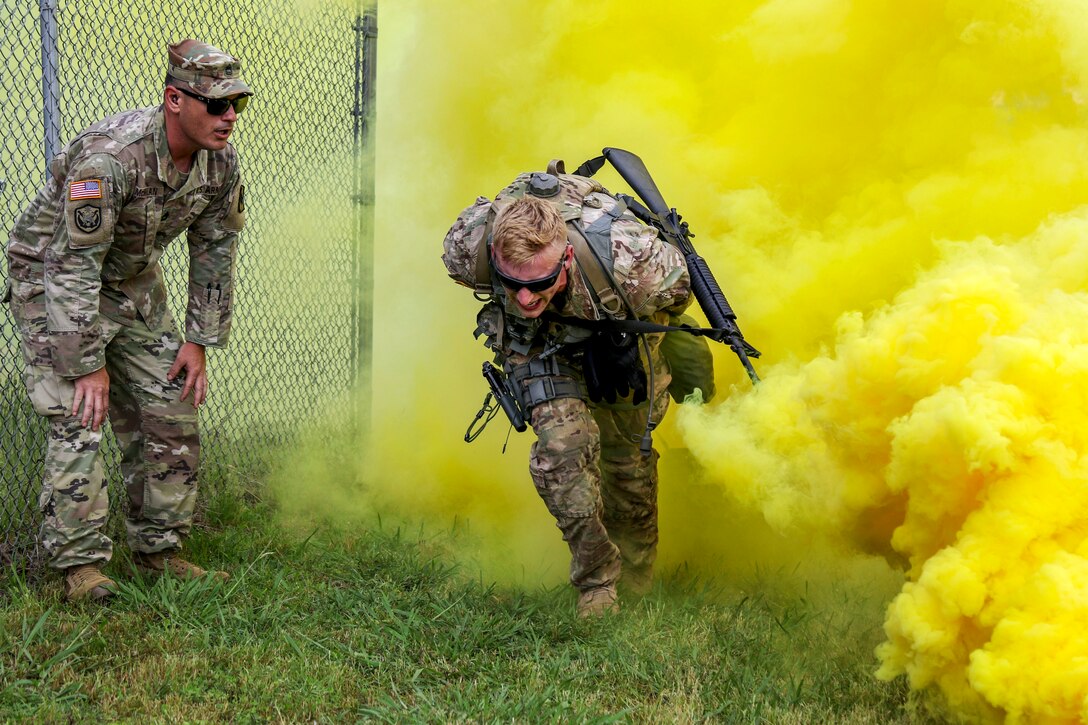 A soldier crouches and runs in front of a thick cloud of yellow smoke, as another observes.