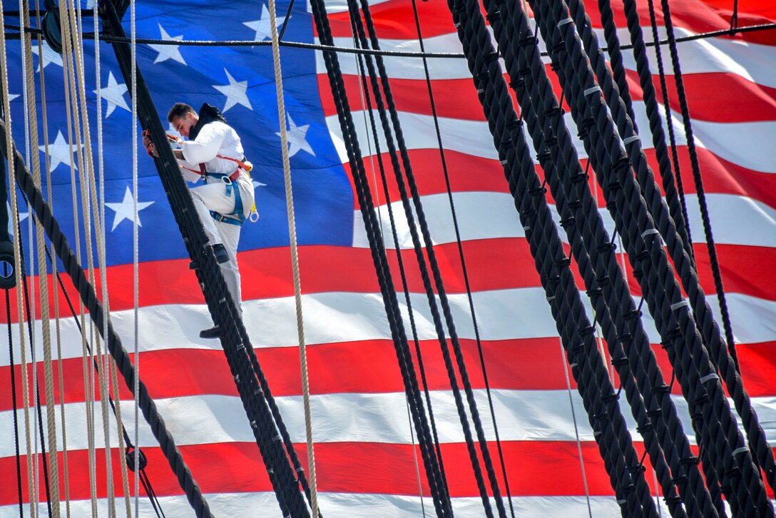 A sailor climbs a rope aboard a ship, with a giant American flag as the backdrop.