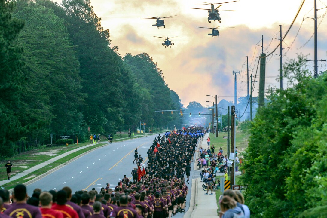 Thousands of soldiers run on a street, as four helicopters fly overhead.