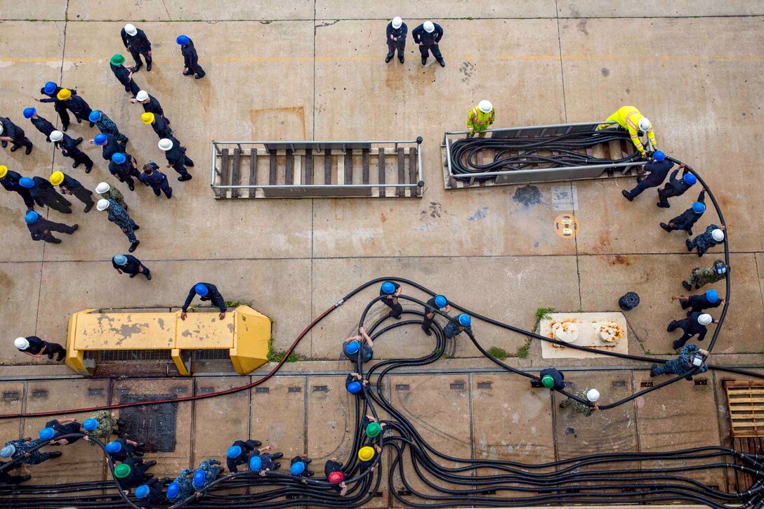 Sailors, shown from overhead, move snaking black lines into a large container on a pier.