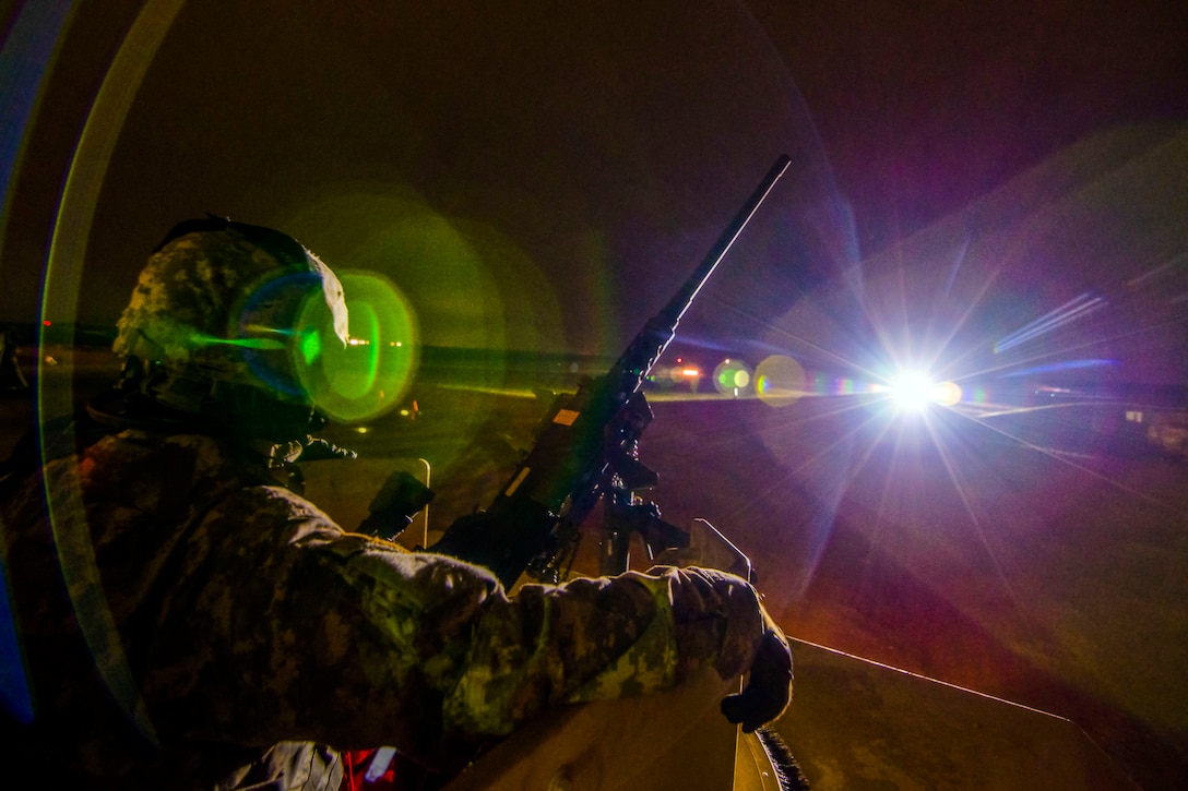 A soldier, shown in profile, sits in a turret at night by a machine gun, illuminated by flashes of light.