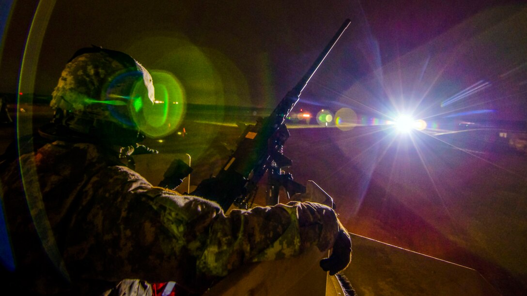 A soldier, shown in profile, sits in a turret at night by a machine gun, illuminated by flashes of light.