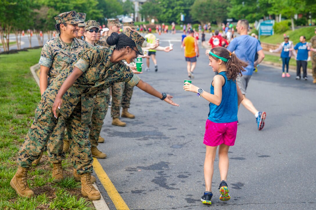 A Marine standing on the sidelines of a road holds her hand to a runner who has just accepted a cup of water from her.