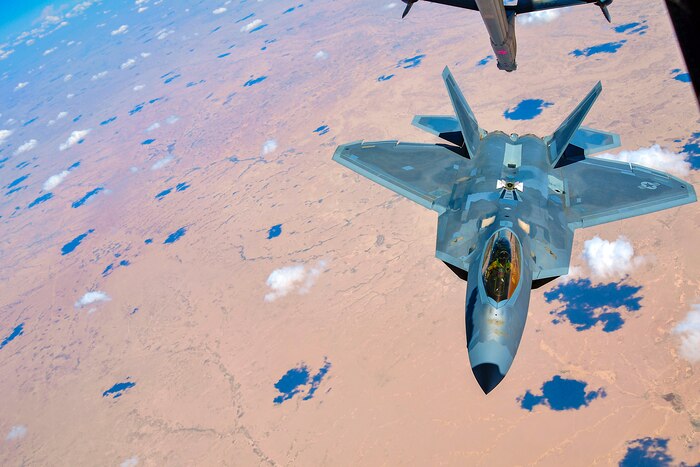 An aircraft boom lowers toward an F-22 Raptor flying over brown terrain, dotted with blue shadows from clouds overhead.