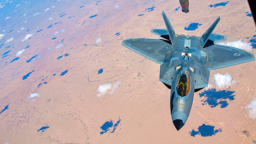 An aircraft boom lowers toward an F-22 Raptor flying over brown terrain, dotted with blue shadows from clouds overhead.