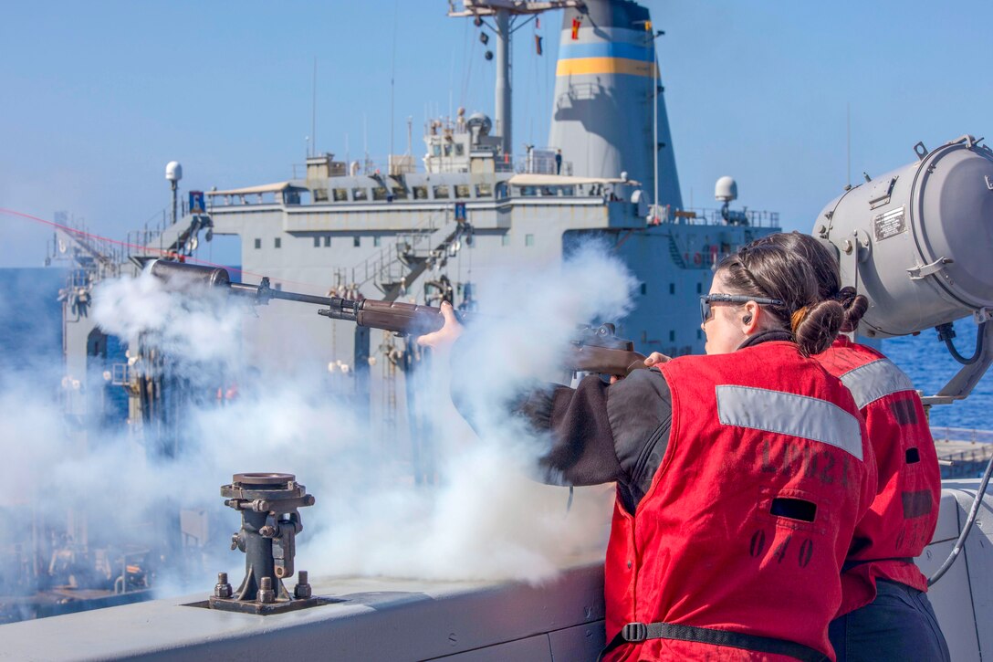 A sailor standing with another sailor, both in red, fires a red line from a gun on a ship's deck, creating a cloud of smoke.