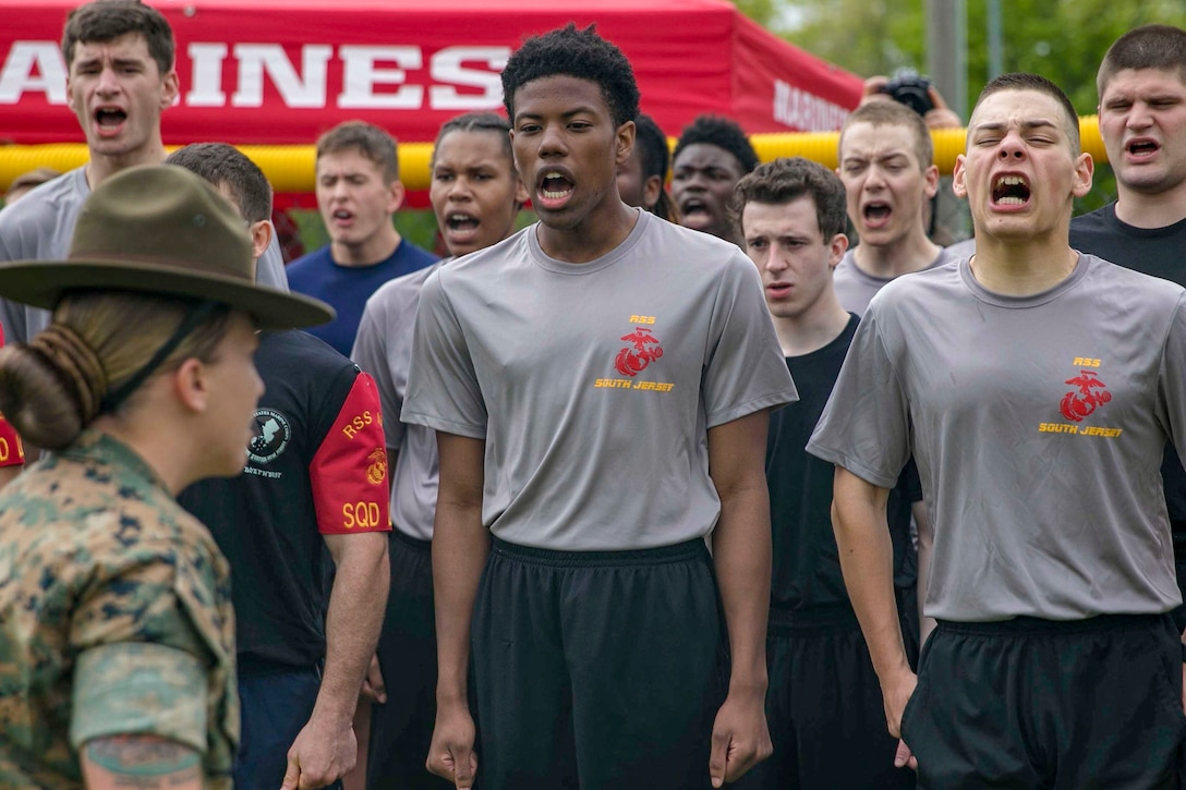 Rows of Marine Corps enlistees shout as a drill sergeant observes.