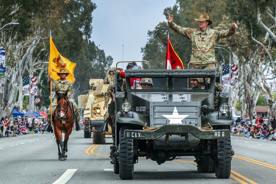 A soldier waves from a military vehicle driving down a parade route.