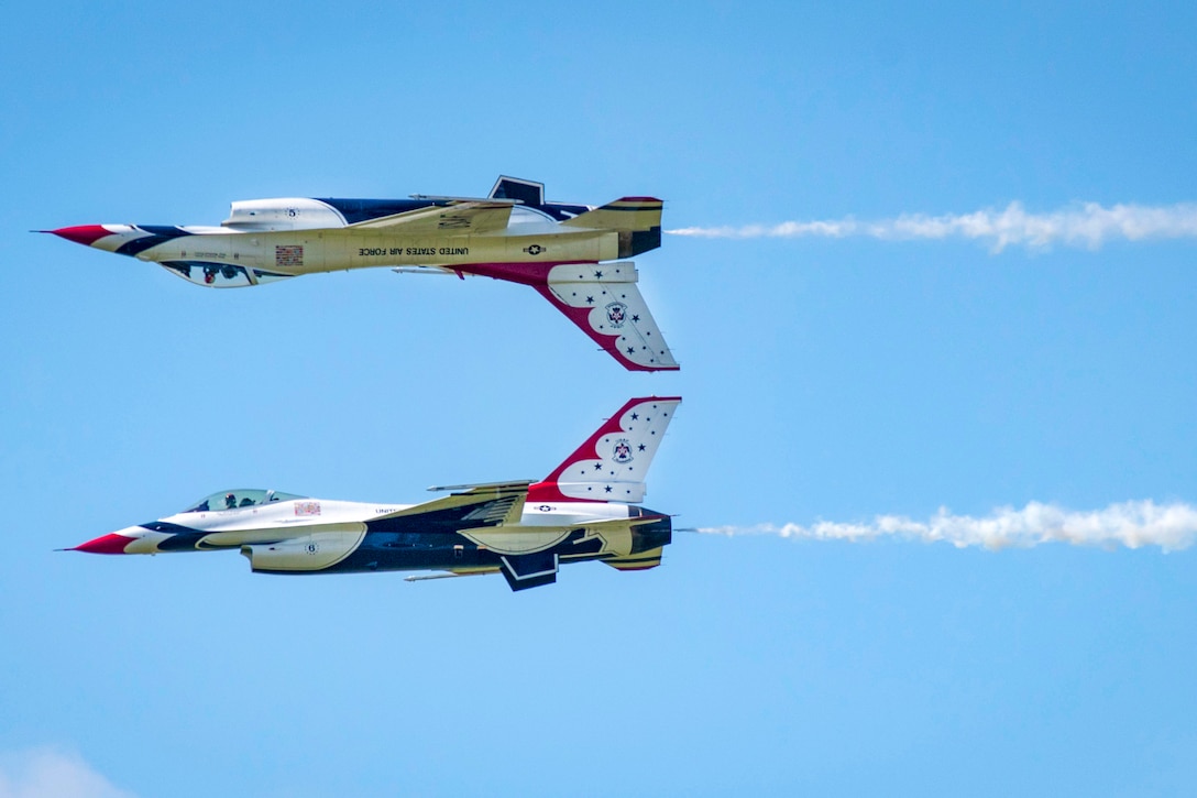 One red, white and blue jet flies upside-down above another in blue skies.
