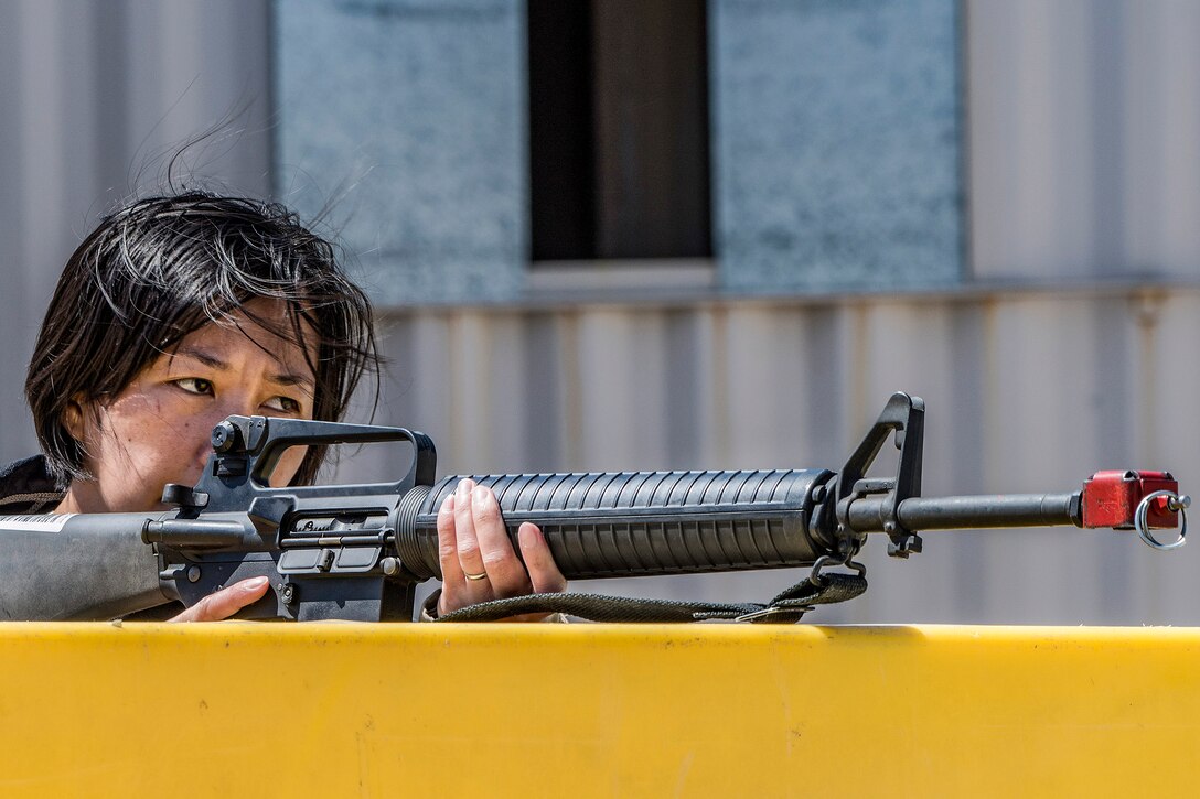 An airman points a rifle while behind a yellow barrier outside.