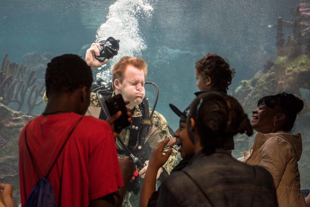 Navy diver plays with children through aquarium glass.