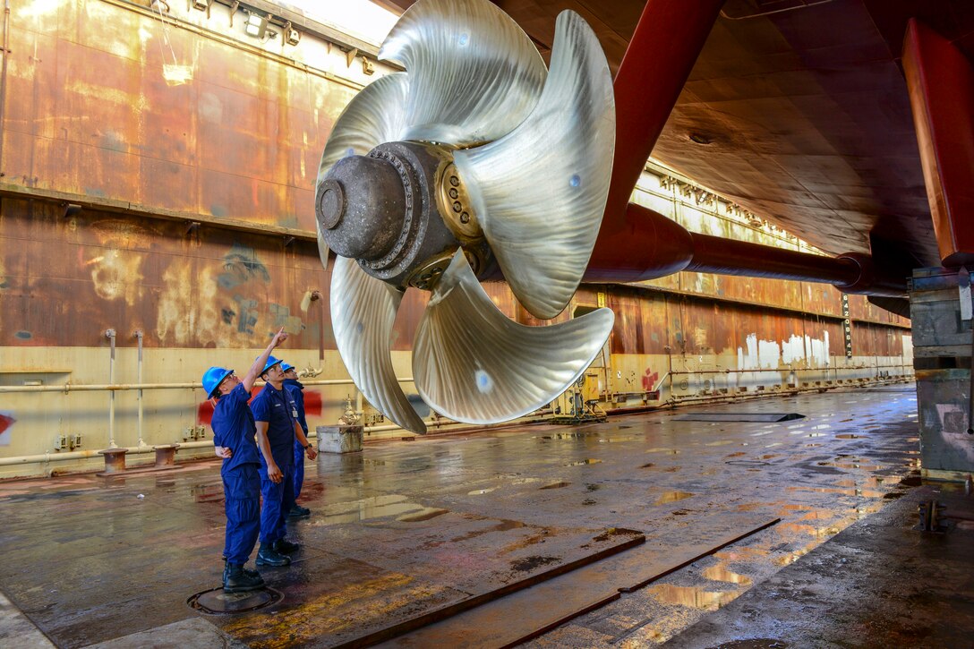 Coast Guard personnel look up at a huge shiny metal propeller.