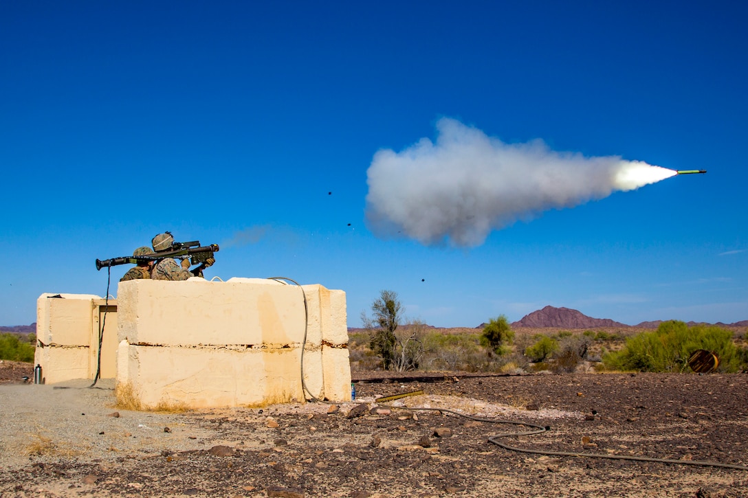 A missile flies low over desert terrain through blue sky, trailed by flames and smoke, as Marines stand behind it wielding a weapon behind a cement-like buttress.