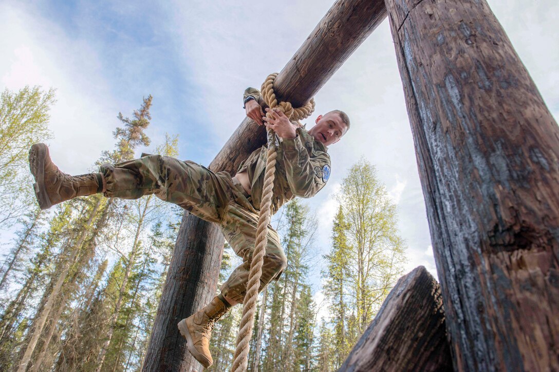 A soldier swings to one side with his legs bent out at different angles while hanging from a rope on a wood obstacle.