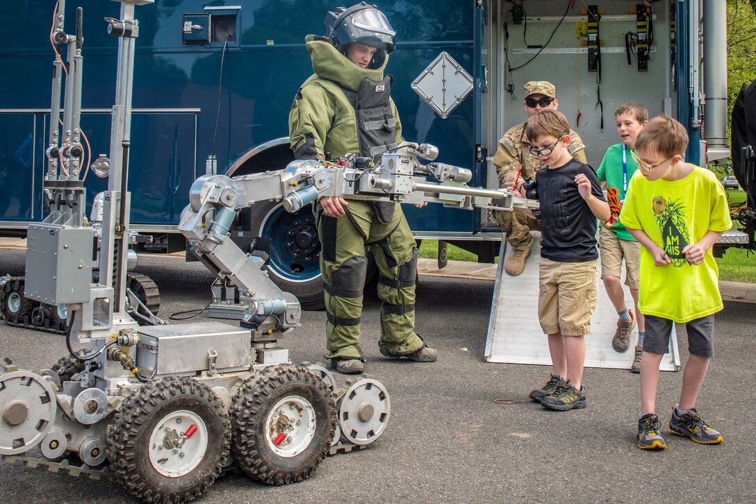 Young students look at a wheeled machine as an airman in a protective bodysuit and helmet stands nearby outside by a trailer.