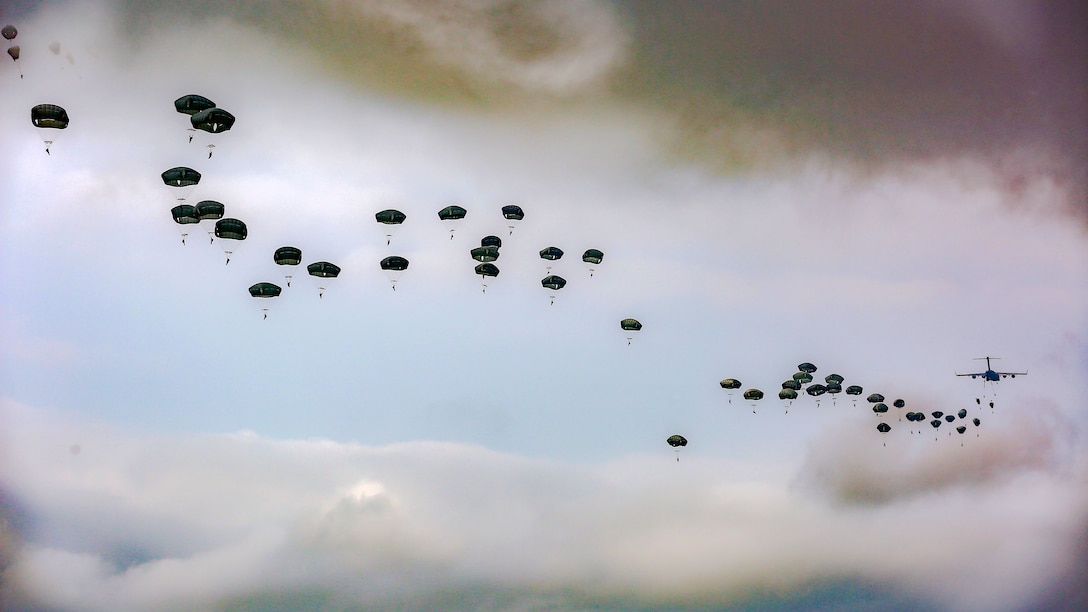 Black parachutes trail an aircraft, dotting a light blue sky with swirling grey clouds.