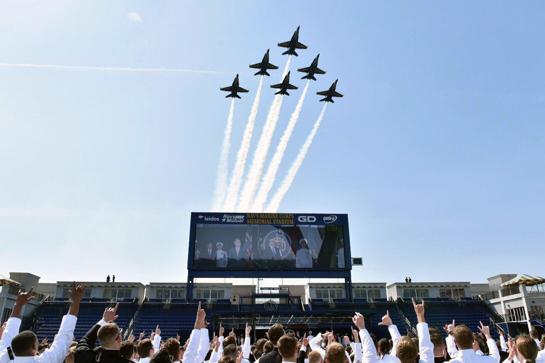 Aircraft fly over a stadium with a crowd.