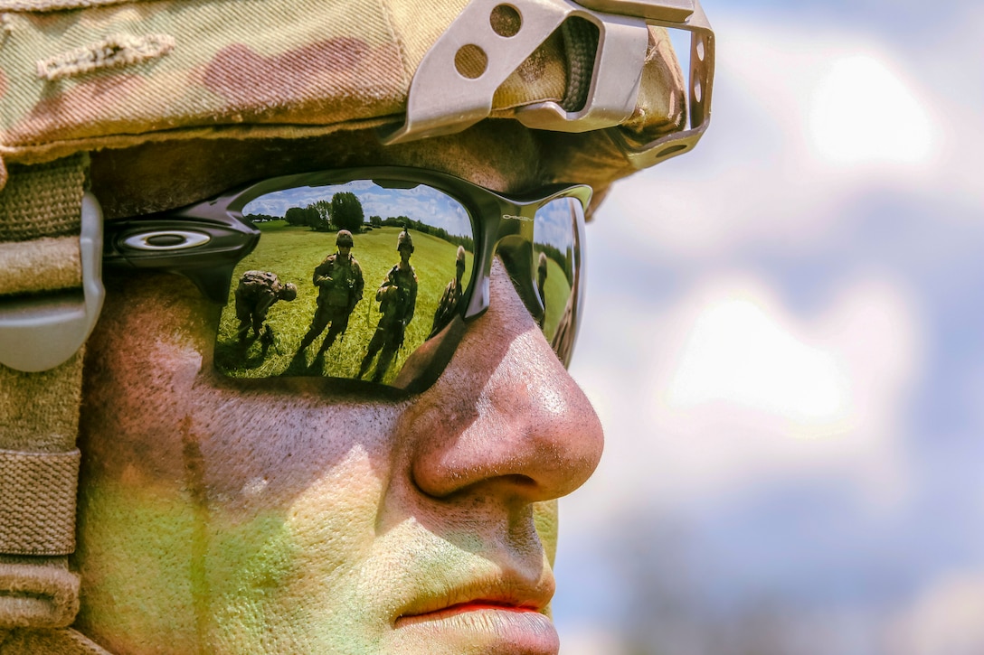 Troops on green grass are reflected in the sunglasses of a soldier staring ahead.