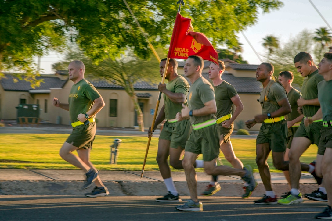 Marines in olive running gear run on a street, as one holds up a red unit guidon.