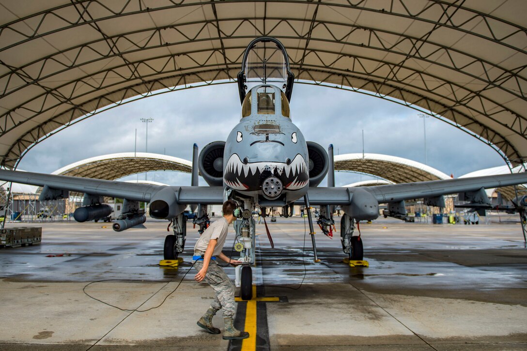 An airman crouches while walking past a hangar with an aircraft with an angry face painted on its nose.