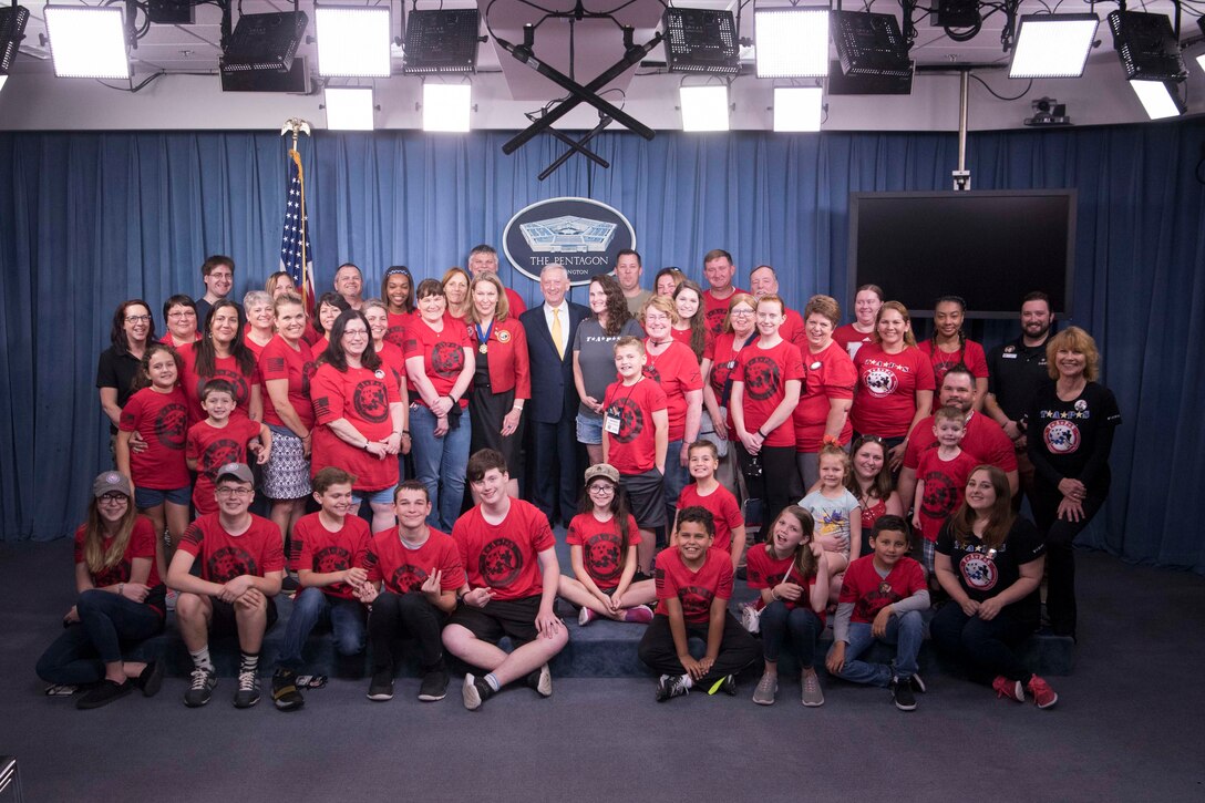 Defense Secretary James N. Mattis stands for a photo with a group of people in matching red shirts in the Pentagon briefing room.