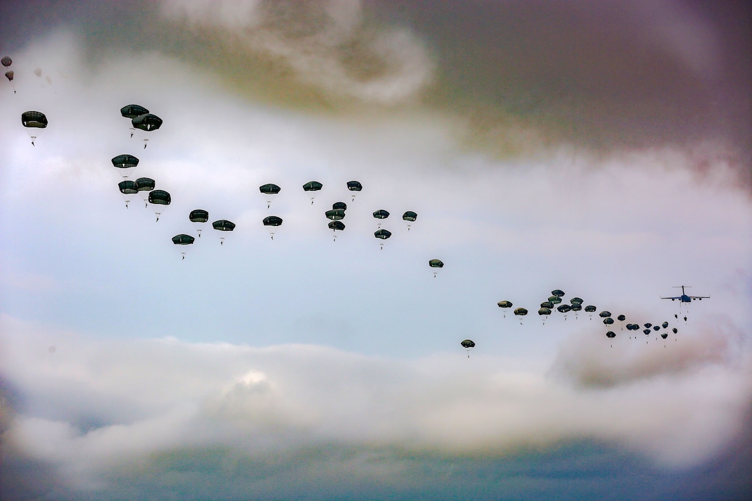 Black parachutes trail an aircraft, dotting a light blue sky with swirling grey clouds.