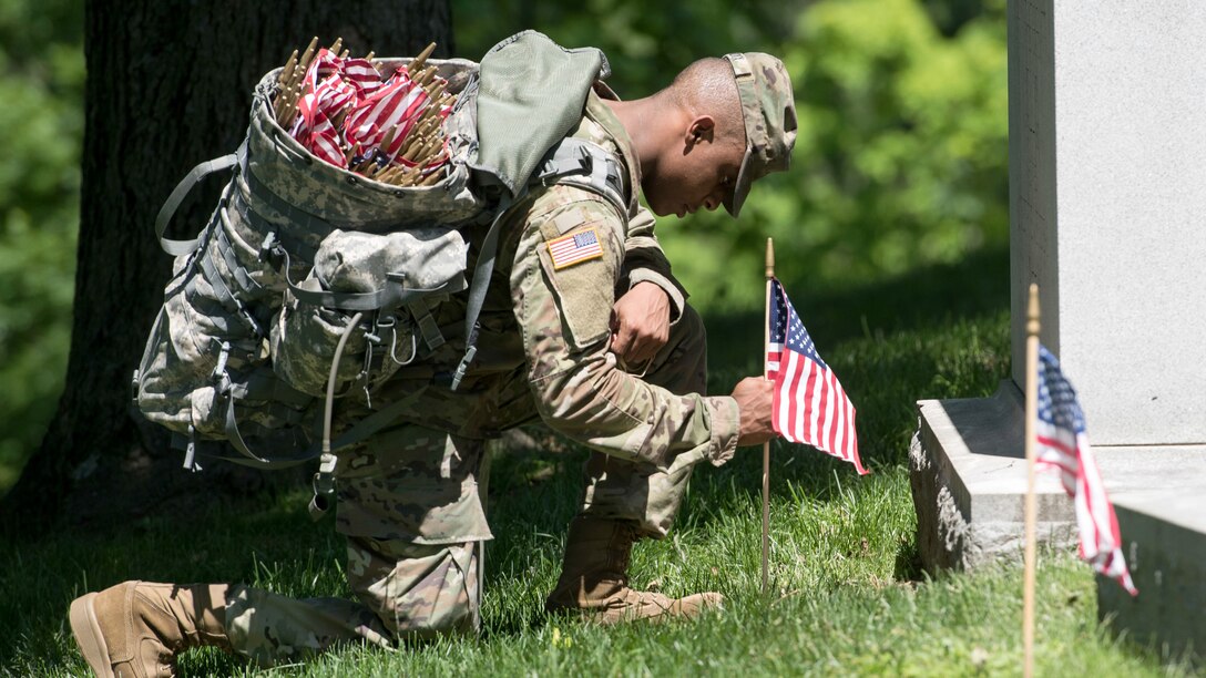 A soldier kneels at a grave to place a flag.