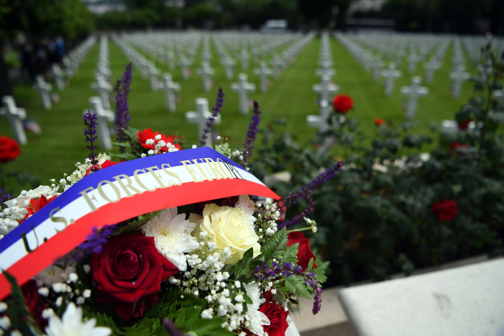 A flower wreath lays on a bench with the Suresnes American Cemetery in the background.