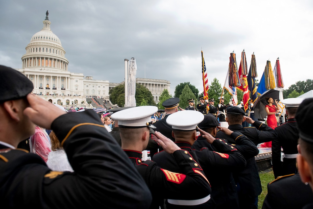 A woman on a stage sings in front of the Capitol building while service members salute.