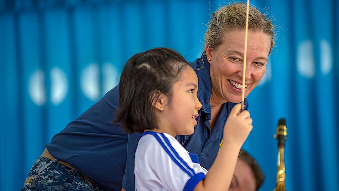 A little girl holds up a conductor's baton as a sailor looks on, framed by a bright blue background.