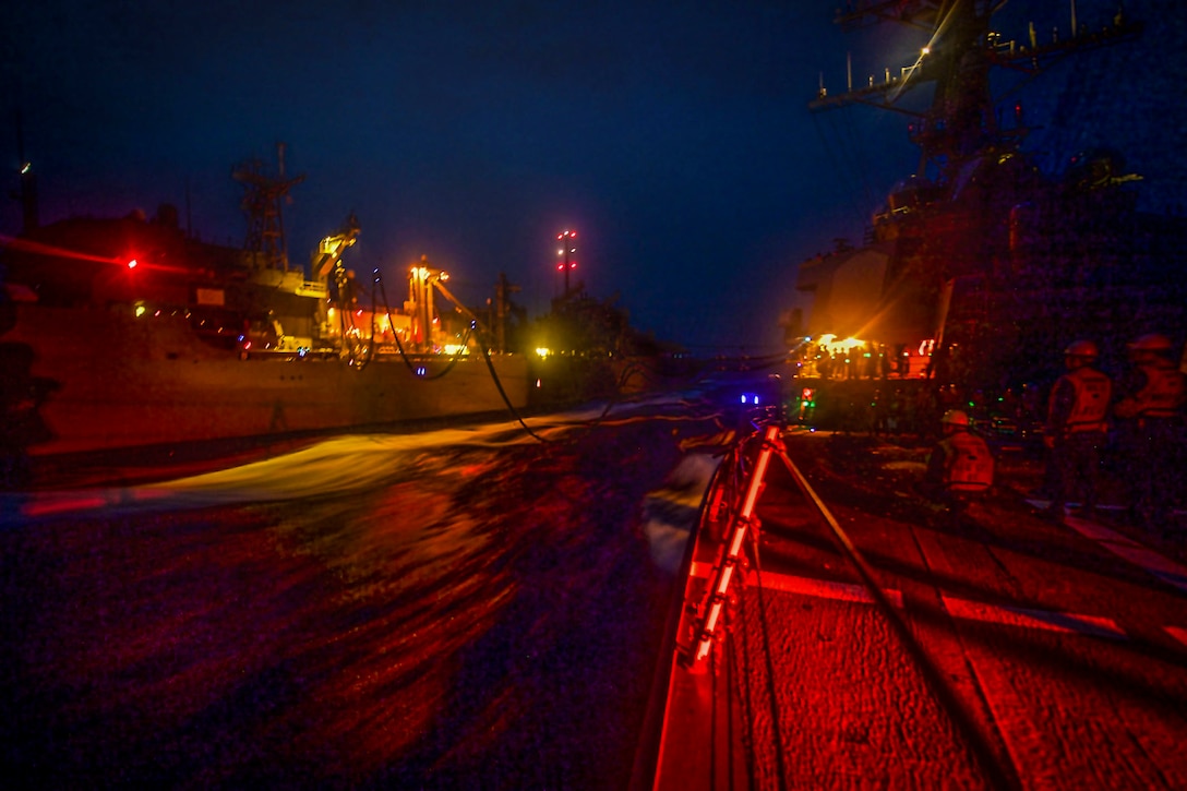 Two ships, illuminated by yellow and red lights, sail side-by-side against a dark blue sky.